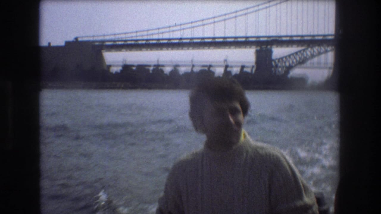 A man in a light knitted sweater sits or stands aboard a moving boat in metadata-supported 1975 Boston, with open water and a large bridge visible behind him.