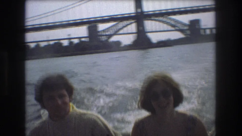 A man and a woman ride on a moving boat near a large bridge, with white wake behind them in metadata-supported Boston home-movie footage from 1975.