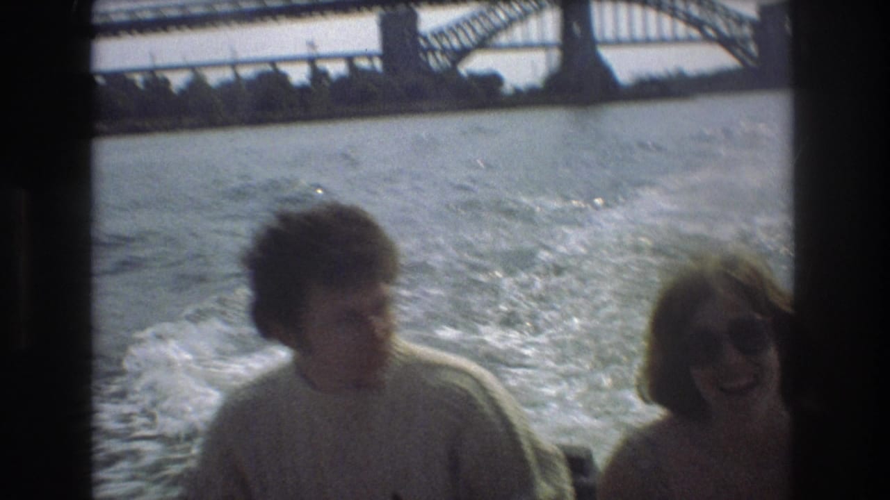 Two adults appear in soft-focus 8mm footage on a moving boat, the woman smiling in sunglasses as bridge structure and water fill the background.