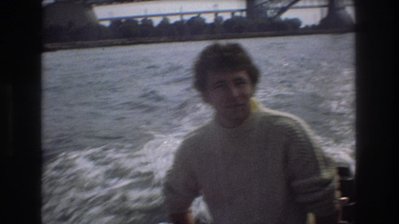 A man in a pale sweater is framed on the right side of a moving boat shot, with foamy wake, open water, and partial bridge structure behind him.