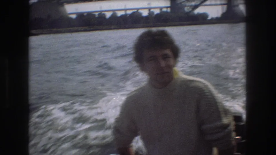 A man in a pale sweater is framed on the right side of a moving boat shot, with foamy wake, open water, and partial bridge structure behind him.