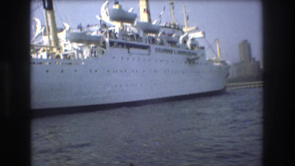 Large white passenger ship seen from the water in Boston in 1975, with upper decks, rigging, harbor water, and distant waterfront structures visible through soft 8mm focus.