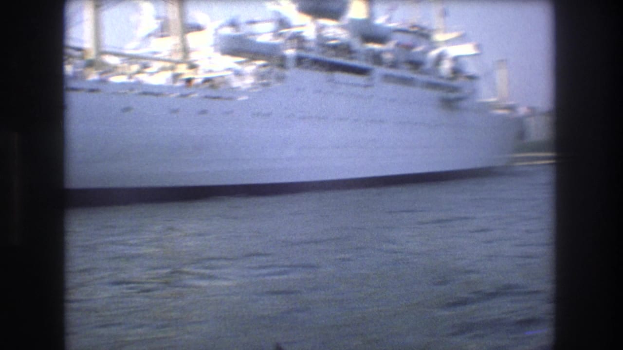 Motion-blurred 1975 Boston harbor frame of a large white passenger ship with its hull and upper deck passing across rippled water, with faint waterfront structures at the edge of the frame.