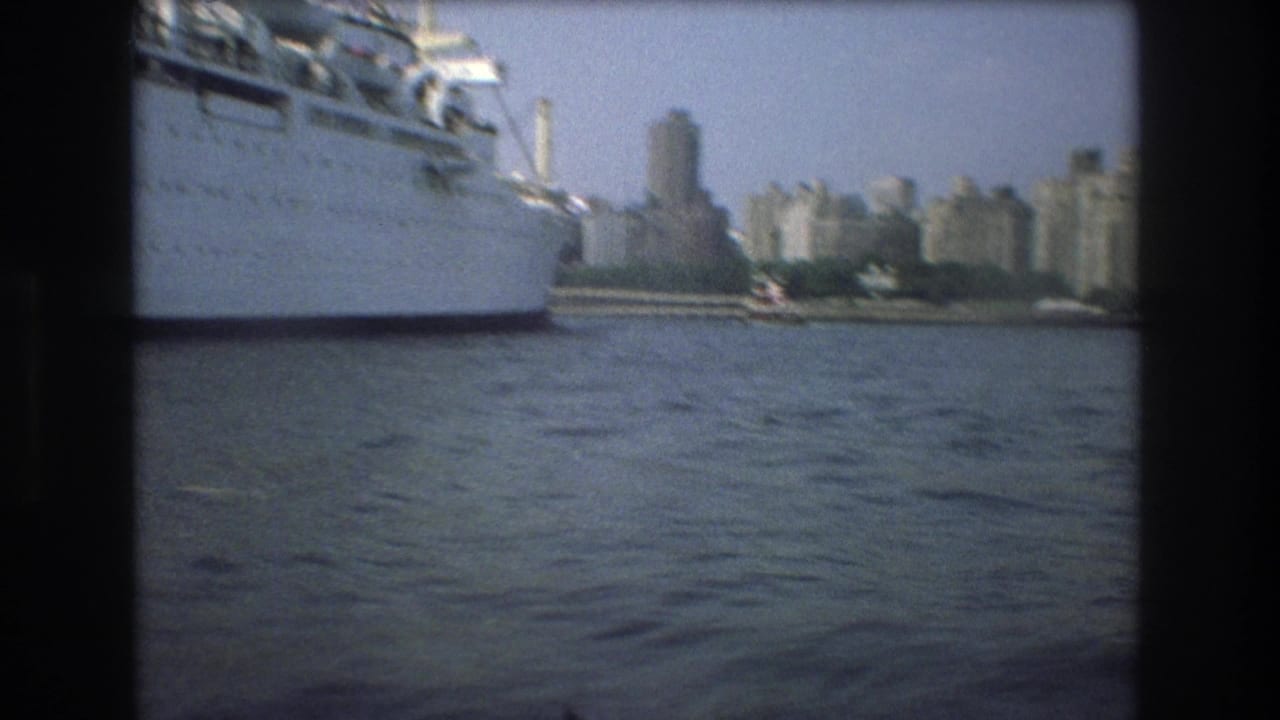 1975 8mm frame showing a large white passenger ship at left with a distant urban skyline across the water.