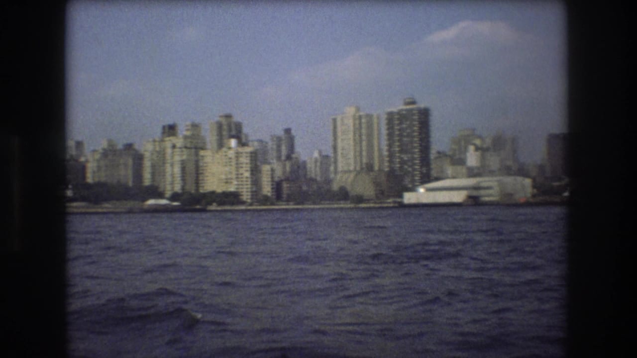 1975 8mm frame of waterfront high-rises and a low white building viewed across choppy water from a moving boat.