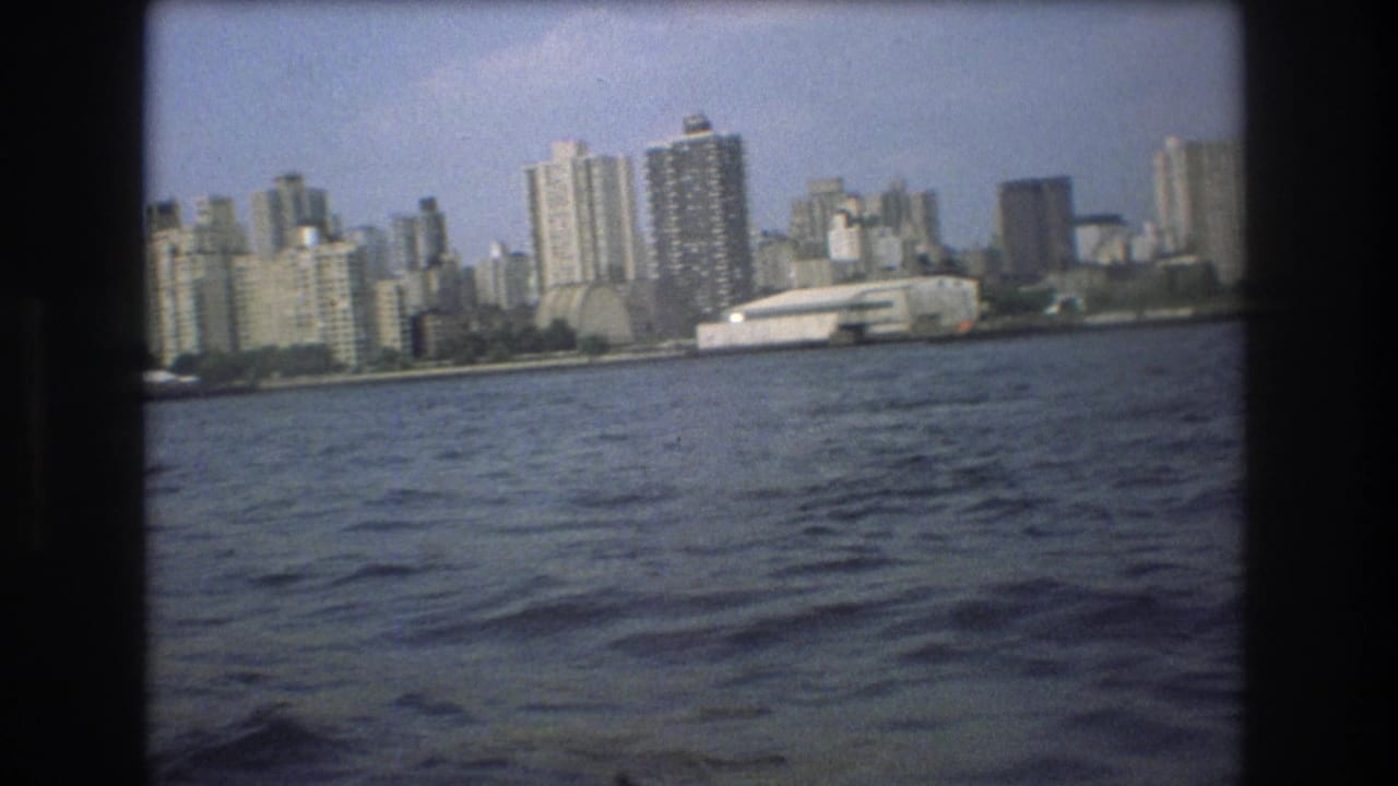 1975 8mm frame with a slightly tilted view of rippling water, waterfront buildings, and a low white structure along the shore.