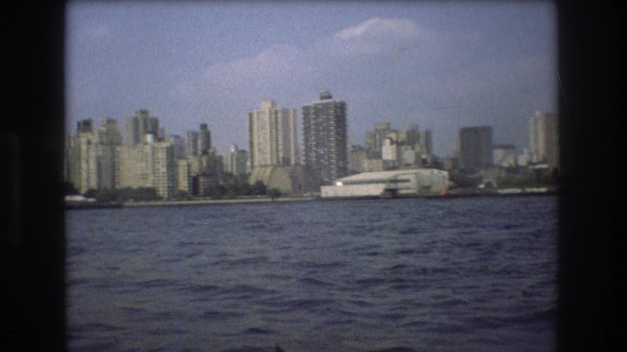 1975 8mm home-movie view of Boston seen from the water, with high-rise buildings and a large white waterfront structure beyond rippled harbor water.