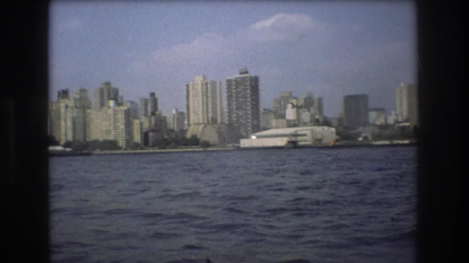 1975 8mm home-movie view of Boston seen from the water, with high-rise buildings and a large white waterfront structure beyond rippled harbor water.
