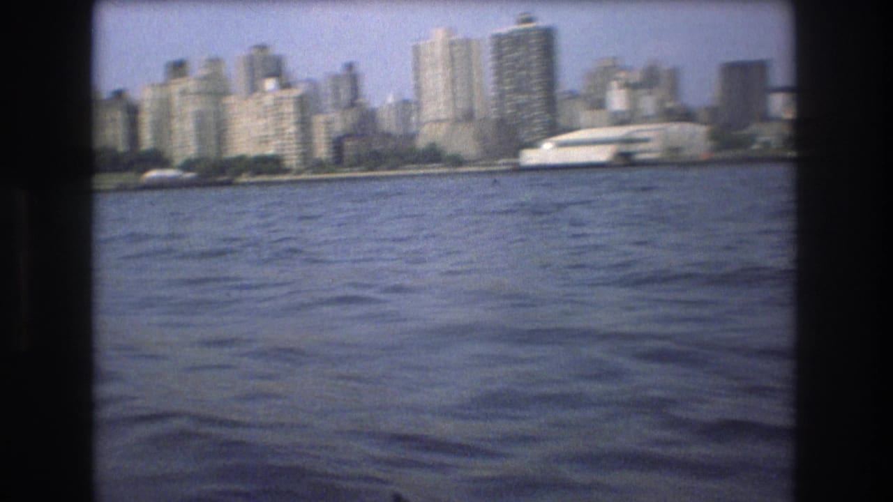 Soft 8mm frame of the Boston waterfront skyline in 1975, showing clustered towers, a low shoreline, and rippled harbor water from a boat-level angle.