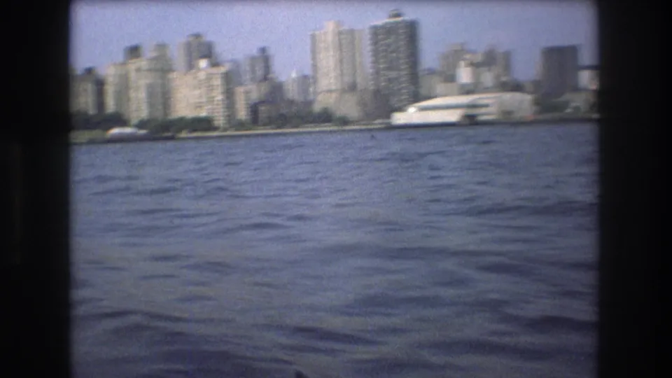 Soft 8mm frame of the Boston waterfront skyline in 1975, showing clustered towers, a low shoreline, and rippled harbor water from a boat-level angle.