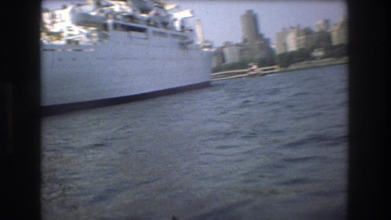 Boat-level 1975 view near Boston with the side of a large white vessel at left, choppy harbor water below, and city towers in the background.