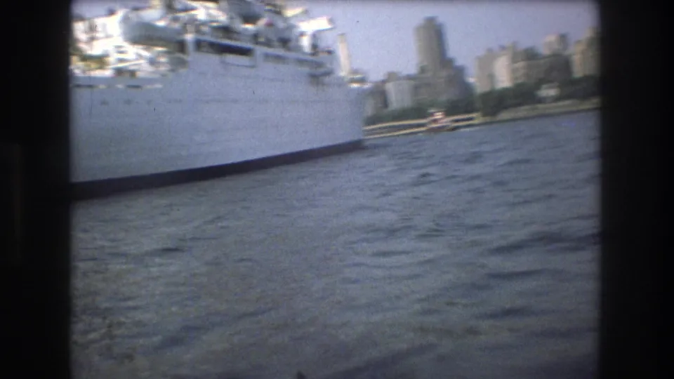 Boat-level 1975 view near Boston with the side of a large white vessel at left, choppy harbor water below, and city towers in the background.