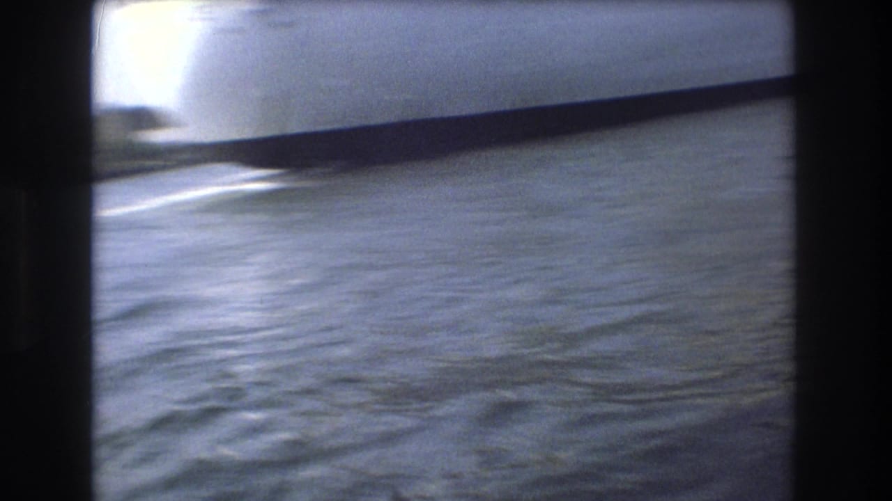 Blue-gray harbor water in Boston, 1975, filmed from a moving boat with a dark 8mm frame edge and a partial vessel or structure crossing the upper frame.