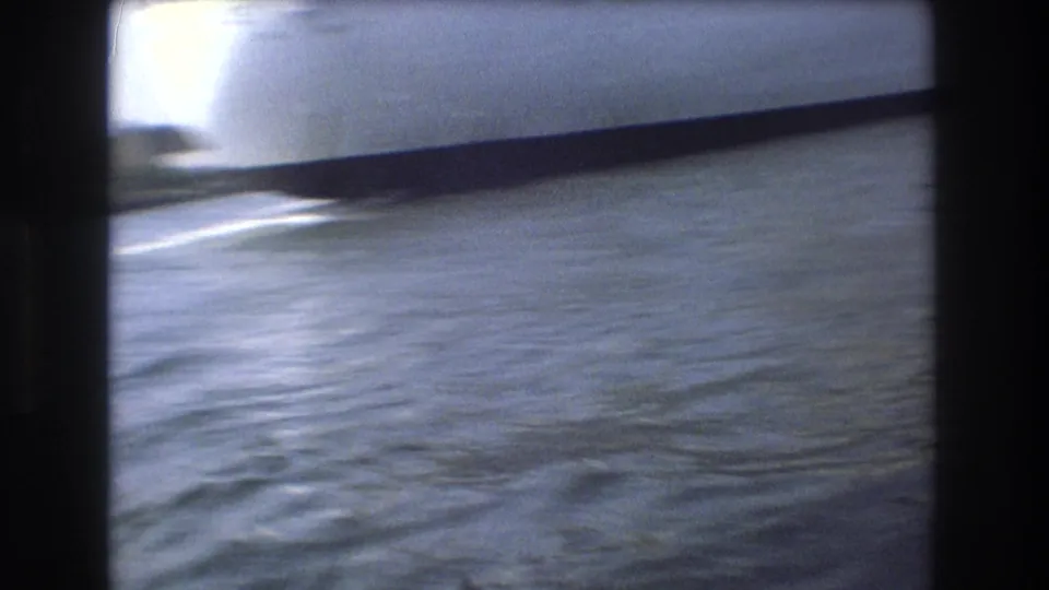 Blue-gray harbor water in Boston, 1975, filmed from a moving boat with a dark 8mm frame edge and a partial vessel or structure crossing the upper frame.