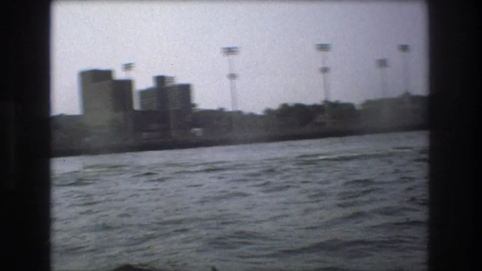 Distant Boston waterfront buildings and tall harbor-side poles appear across choppy water in a soft, grainy 1975 8mm frame.