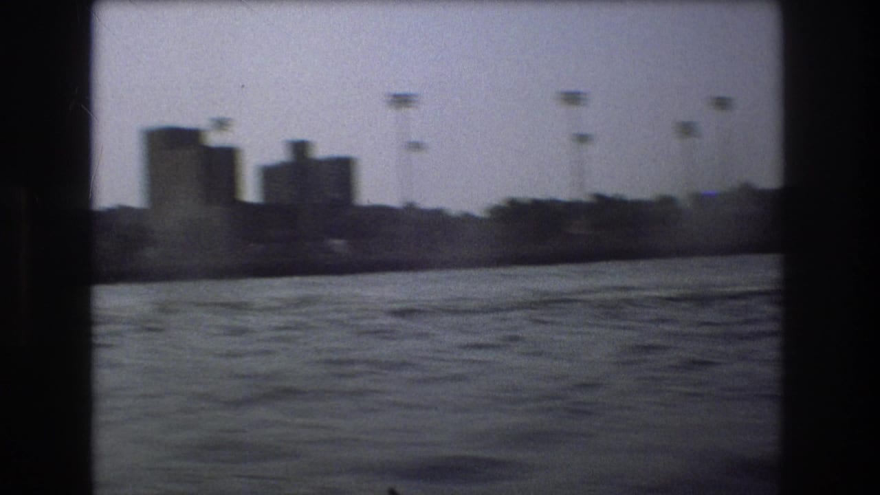 A blocky industrial-looking Boston shoreline is seen across rippled harbor water, with tall poles and dark 8mm frame borders visible.