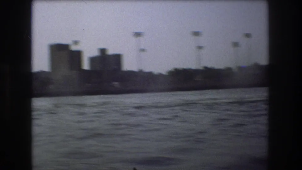A blocky industrial-looking Boston shoreline is seen across rippled harbor water, with tall poles and dark 8mm frame borders visible.