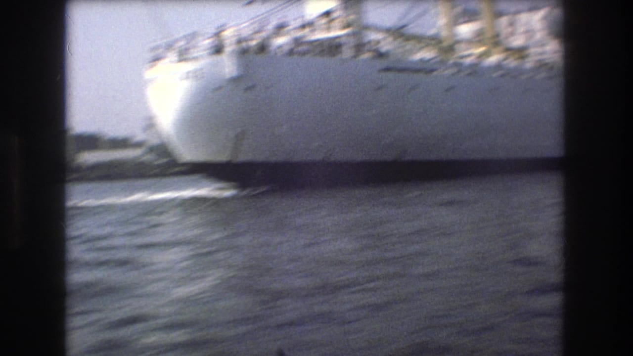 The bow and side of a large white vessel pass close to the camera on choppy Boston harbor water in a grainy 1975 home-movie frame.
