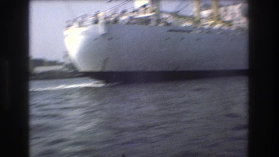 The bow and side of a large white vessel pass close to the camera on choppy Boston harbor water in a grainy 1975 home-movie frame.