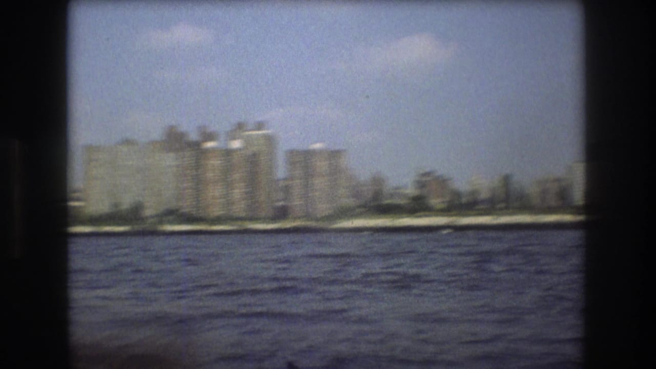 1975 Boston harbor frame showing blocky waterfront buildings and shoreline viewed from a boat across rippled water.