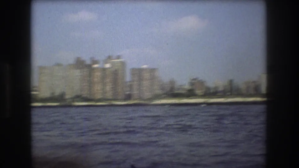 1975 Boston harbor frame showing blocky waterfront buildings and shoreline viewed from a boat across rippled water.