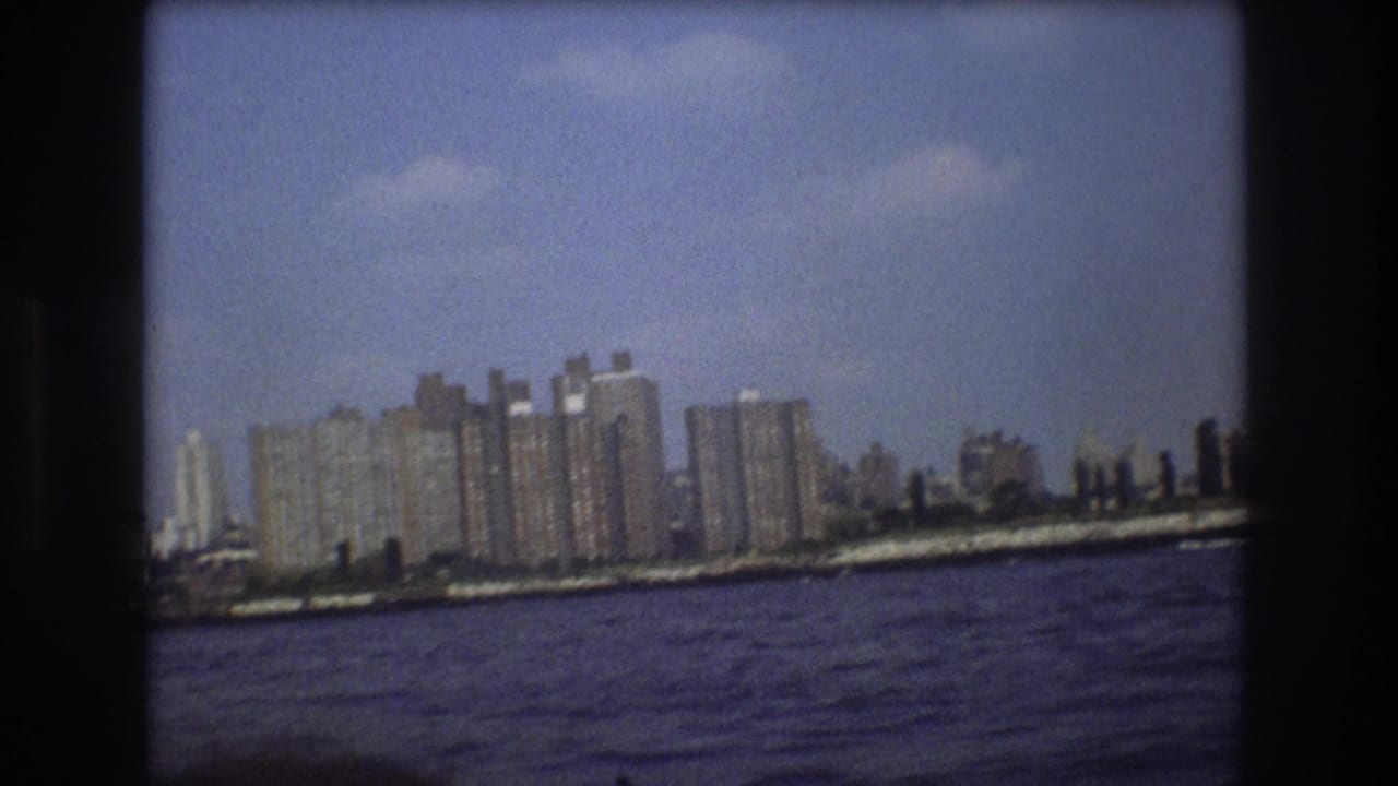 1975 Boston Harbor home-movie frame with tall waterfront buildings, a narrow shoreline, and dark blue water in the foreground.