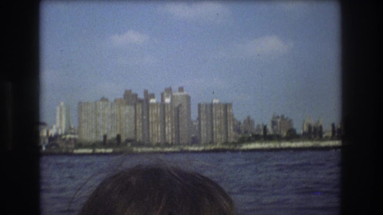1975 Boston home-movie frame showing a distant waterfront skyline across rippling water, with the top of a passenger's head at the lower edge.