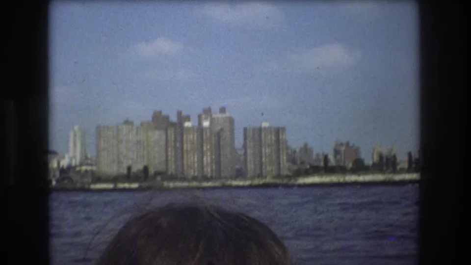 1975 Boston home-movie frame showing a distant waterfront skyline across rippling water, with the top of a passenger's head at the lower edge.