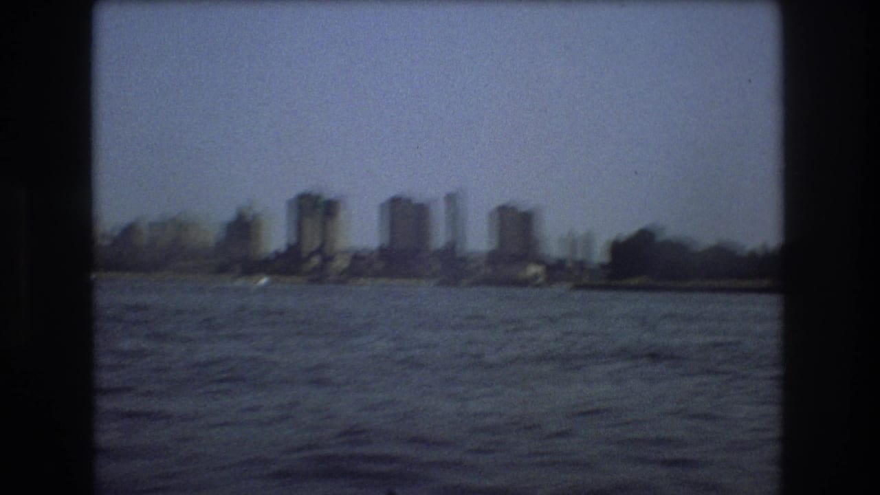 1975 Boston waterfront frame with rippling harbor water, a distant bridge structure, and high-rise buildings along the shoreline.