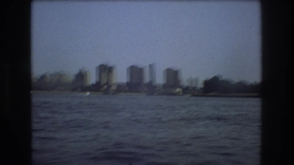 1975 Boston waterfront frame with rippling harbor water, a distant bridge structure, and high-rise buildings along the shoreline.