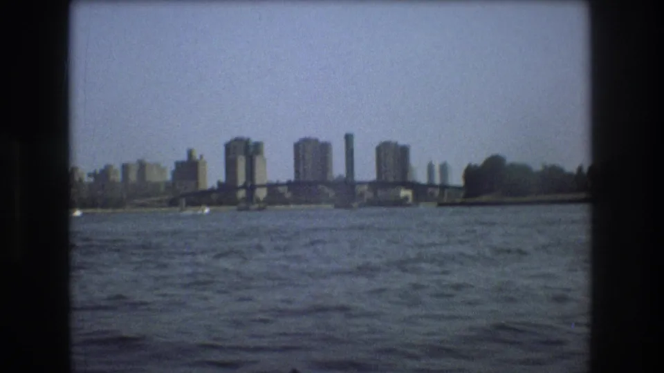 1975 Boston harbor view from a small boat, with open water in the foreground, a low bridge, and distant city buildings along the waterfront.