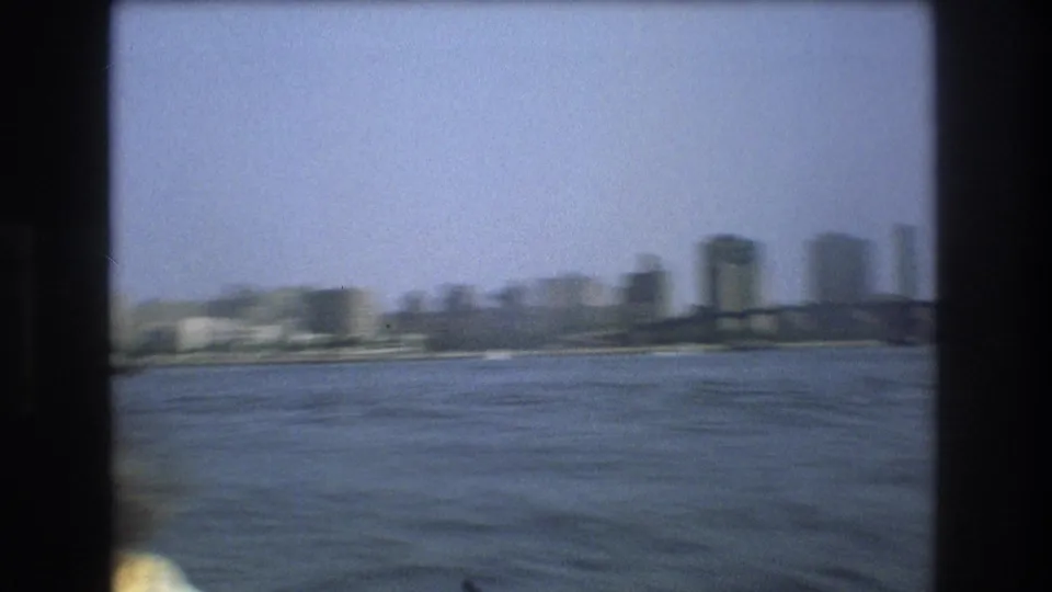 Soft 8mm frame of Boston harbor in 1975, showing water, a bridge, and distant high-rise buildings as the camera moves from a small boat.