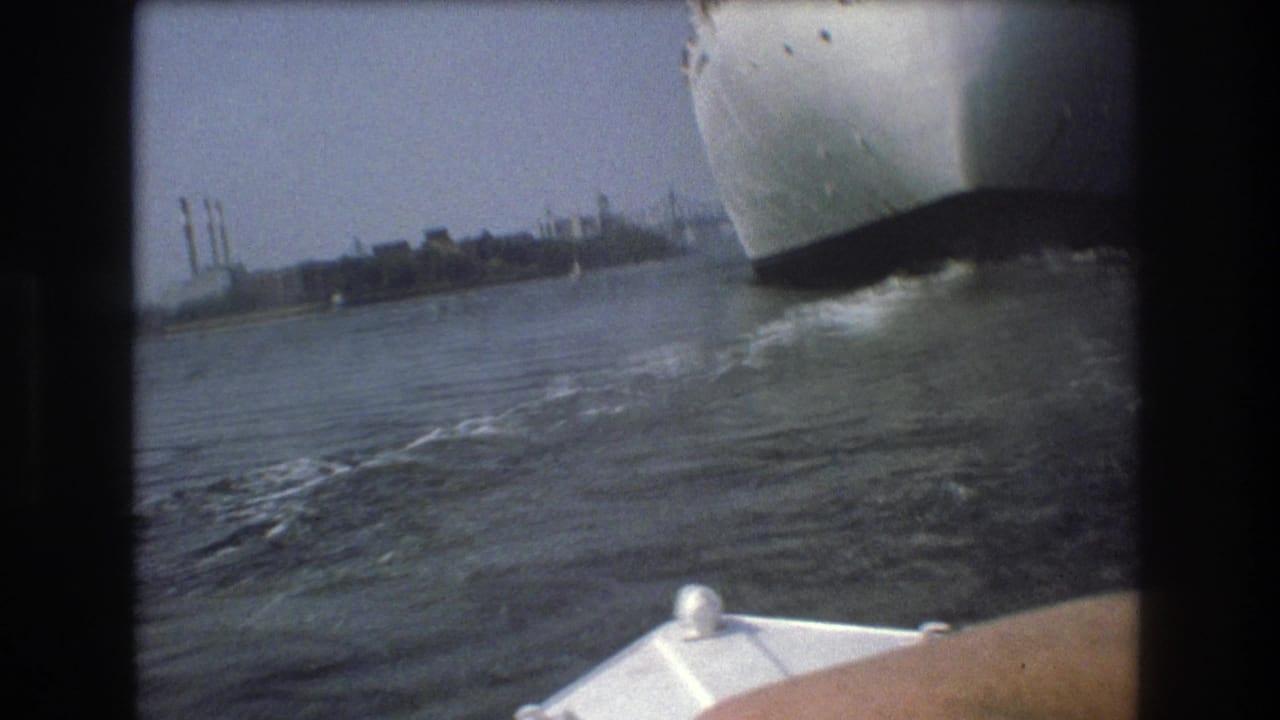 A large white vessel passes close to the camera on Boston harbor in 1975, with the bow of the small filming boat visible at the bottom of the frame.