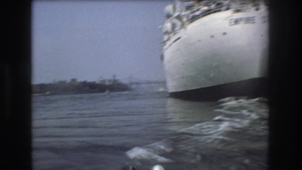 1975 Boston harbor home-movie frame showing the bow of the white Empire State vessel from a nearby boat, with water wake and distant shoreline visible.