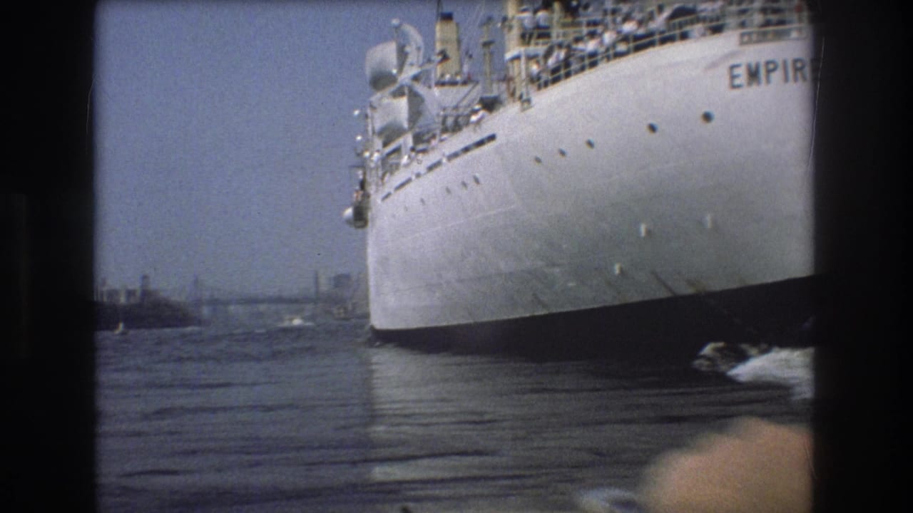 1975 Boston harbor home-movie frame showing the side and bow area of the Empire State vessel, with passengers faintly visible above and a distant bridge or shoreline structure beyond the water.