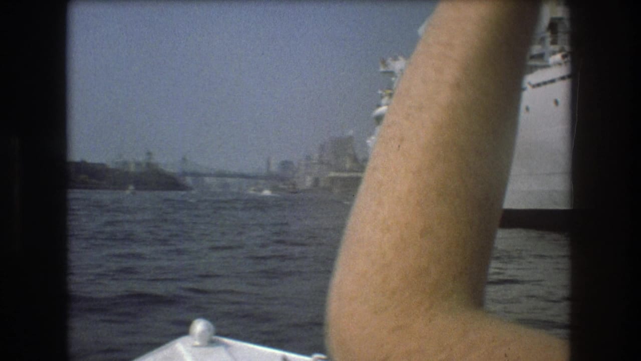 1975 Boston harbor home-movie frame showing a passenger’s raised arm from a small boat, with the white ship and distant waterfront in the background.