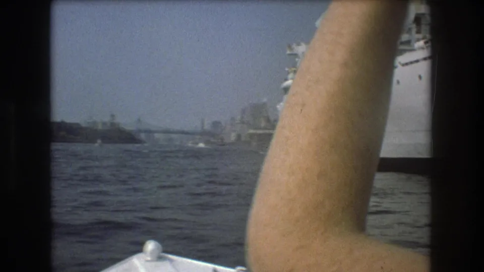 1975 Boston harbor home-movie frame showing a passenger’s raised arm from a small boat, with the white ship and distant waterfront in the background.