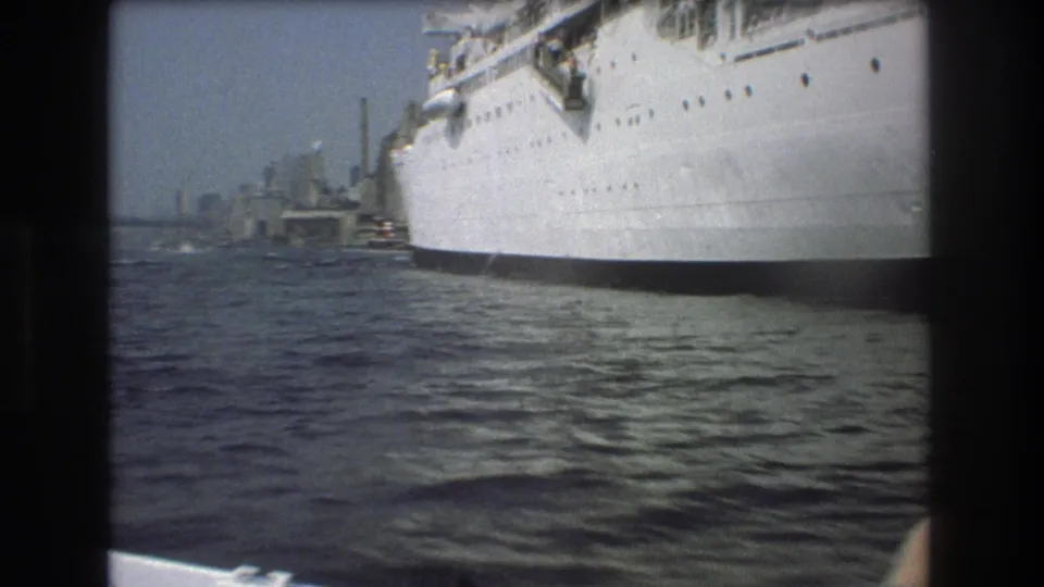1975 Boston home-movie frame showing a large white passenger ship close to the camera, choppy harbor water, and distant port structures.