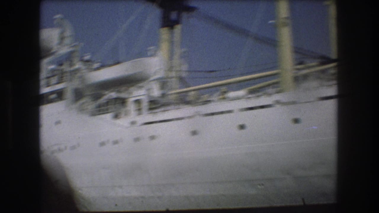 1975 Boston home-movie frame showing a blurred close pass along the white ship, with yellow funnel details and part of a foreground viewer visible.