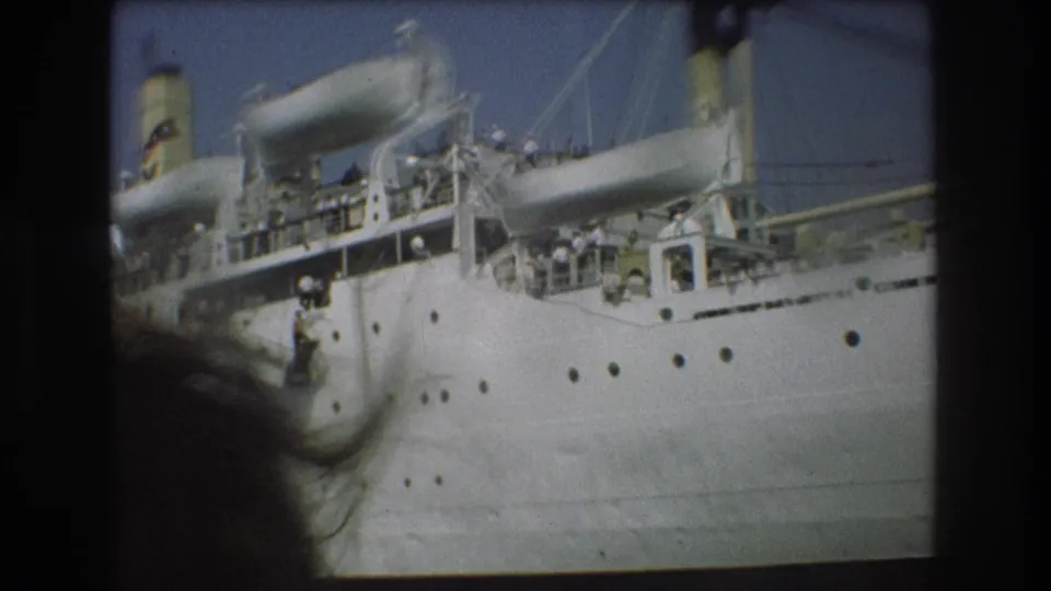 1975 Boston harbor 8mm frame showing a large white passenger ship at a pier, with small figures visible along the decks and dark boat-window framing at the edges.