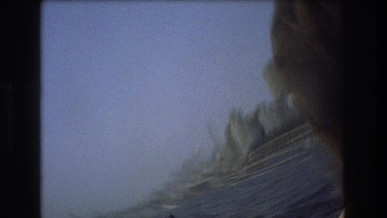 Soft, motion-blurred 8mm view from the moving boat, with a dark foreground edge, pale sky, and indistinct shoreline or waterfront structures beyond.