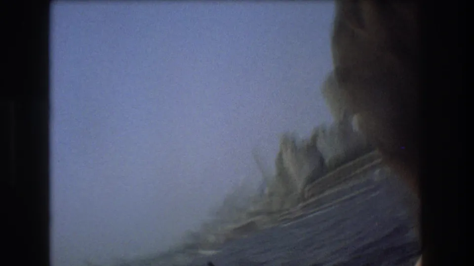 Soft, motion-blurred 8mm view from the moving boat, with a dark foreground edge, pale sky, and indistinct shoreline or waterfront structures beyond.