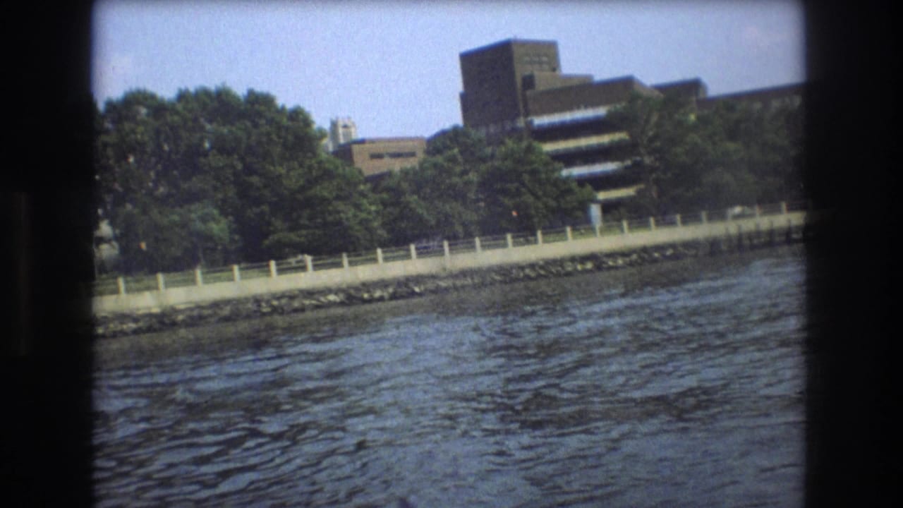 1975 Boston 8mm waterfront view from a small boat, showing rippling water in the foreground, a tree-lined shore, a pale fence, and modern buildings behind it.