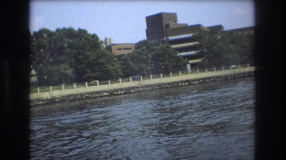 1975 Boston 8mm waterfront view from a small boat, showing rippling water in the foreground, a tree-lined shore, a pale fence, and modern buildings behind it.