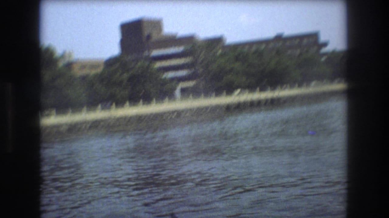 1975 Boston home-movie frame showing a large multi-story building partly obscured by trees beyond a riverside embankment and railing.