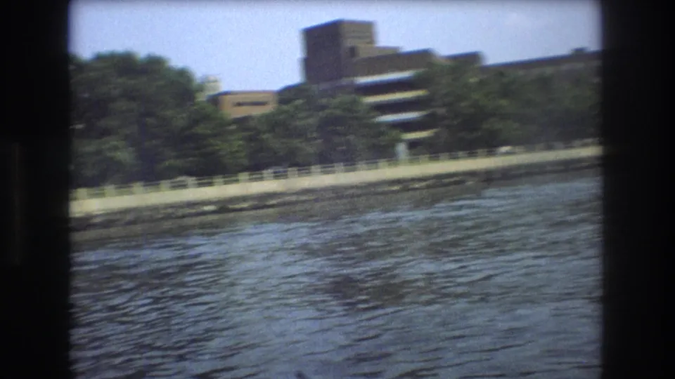 1975 Boston waterfront view from the water, with ripples in the foreground and a large building rising behind trees and a pale railing.