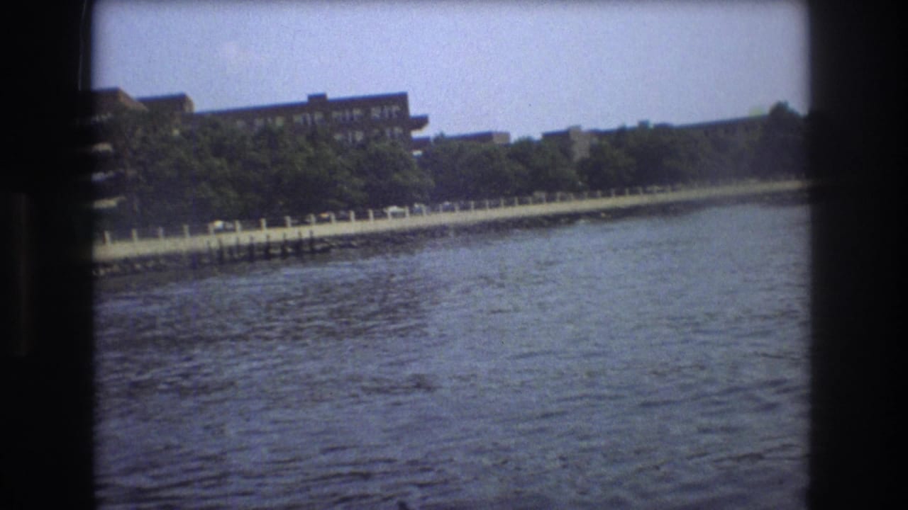 1975 Boston 8mm frame showing water in the foreground, a riverside railing with small parked cars, trees, and multi-story buildings beyond.