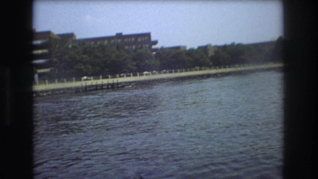 1975 Boston home-movie frame with rippling water, a tree-lined embankment, railing, parked cars, and buildings along the shore.