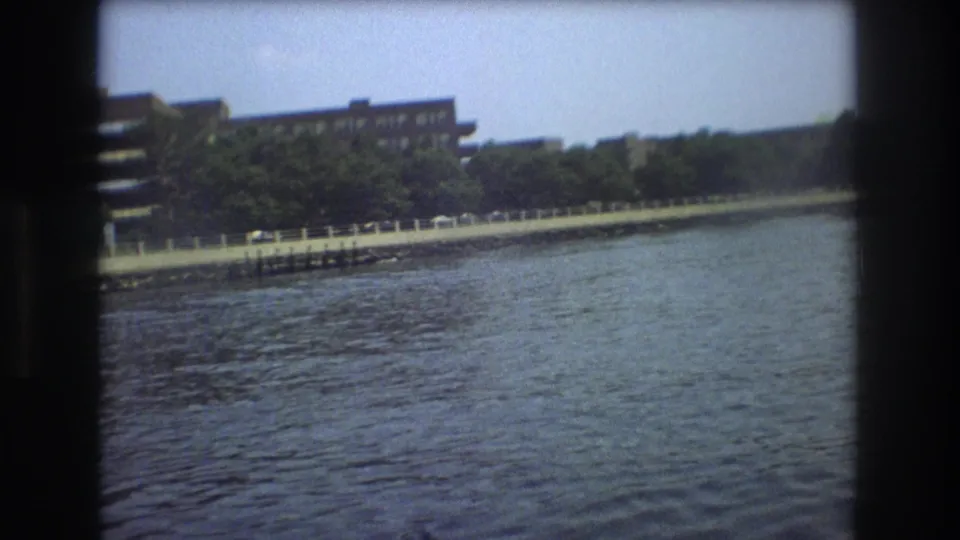 1975 Boston home-movie frame with rippling water, a tree-lined embankment, railing, parked cars, and buildings along the shore.