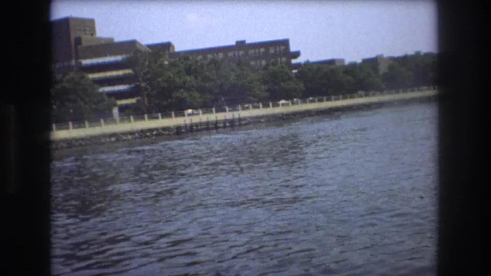 Boston waterfront buildings and a tree-lined shoreline appear across rippling water in 1975 8mm home-movie footage shot from a boat.